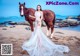 A woman in a wedding dress standing next to a horse on a beach.